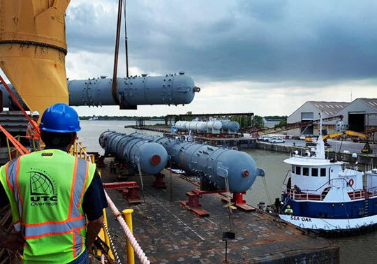UTC Overseas team member in a hard hat and reflective vest on a heavy-lift vessel while a crane transloads oil and gas equipment onto a barge held in place by a tug.